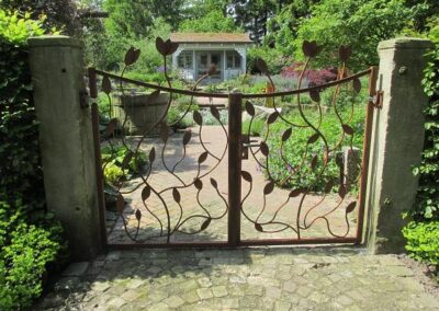 Rustikales Gartentor in Rostoptik mit Unterbogen und augeschmiedeten Rosenblättern. Befestigt mit Halseisenbeschlag an Sandsteinpfosten